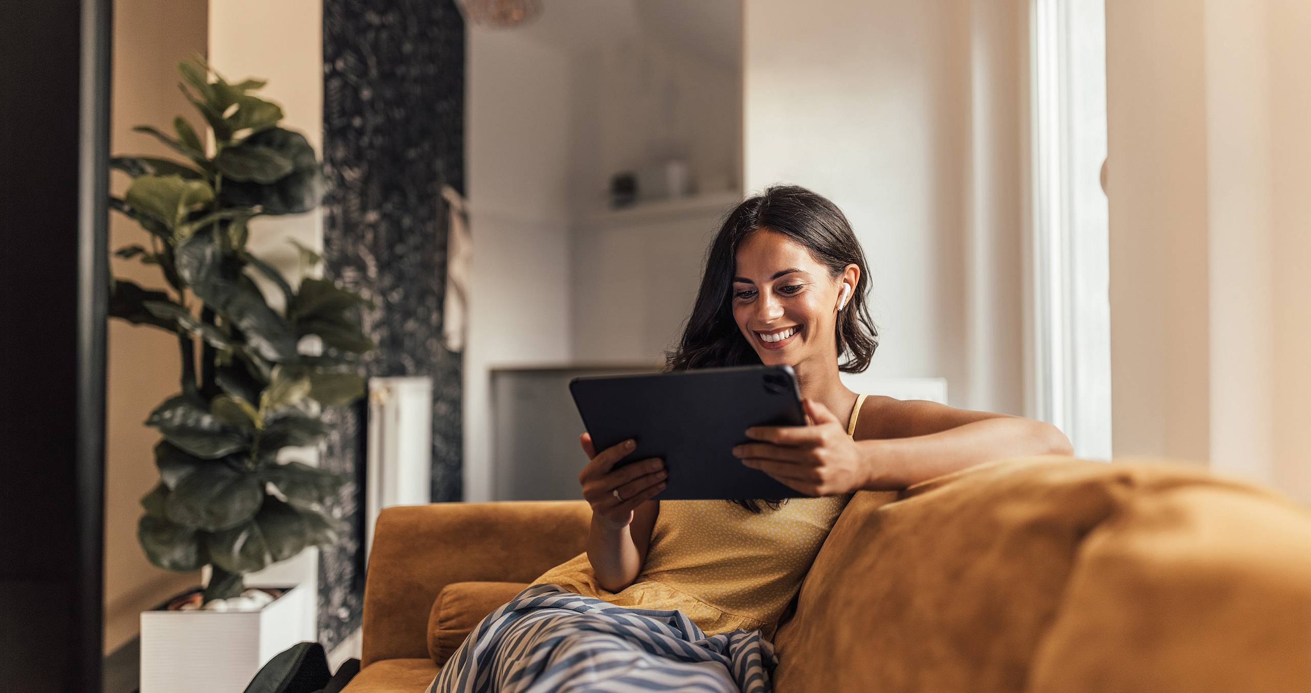 Lächelnde Frau sitzt auf einem Sofa und betrachtet ein Tablet.
