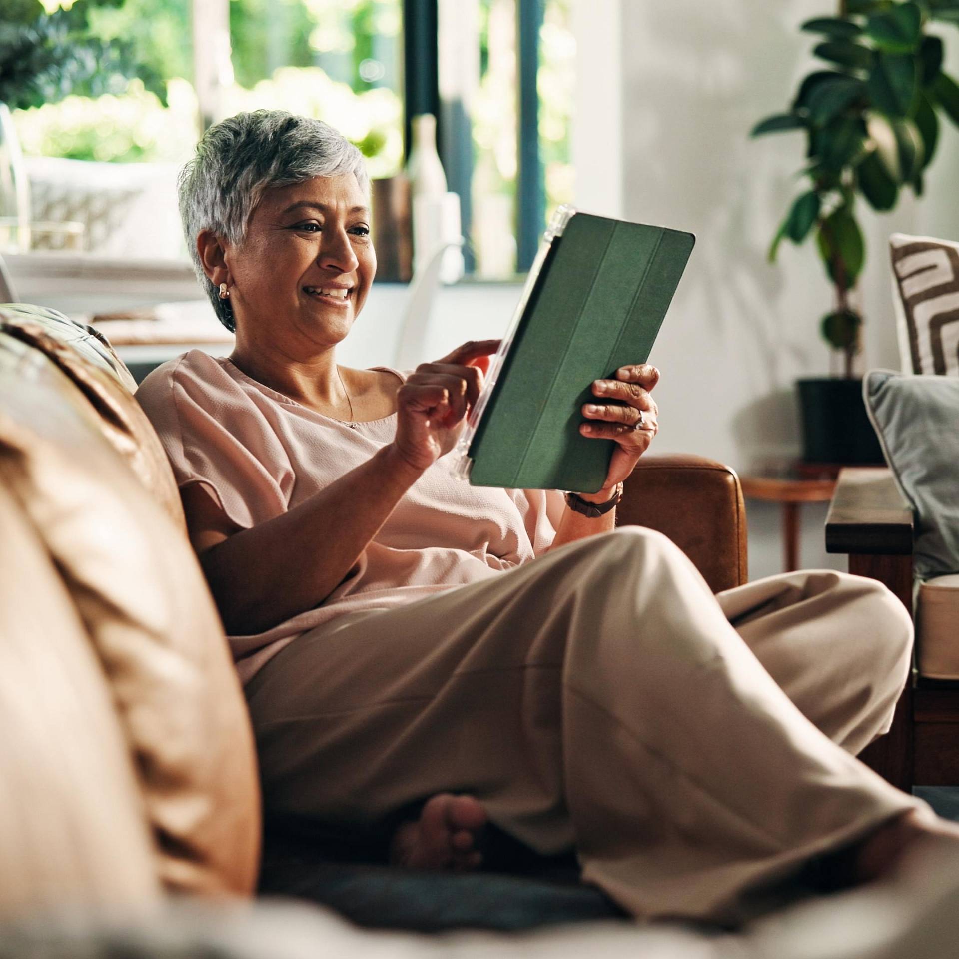 Lächelnde ältere Frau sitzt auf einem Sofa und bedient ein Tablet.