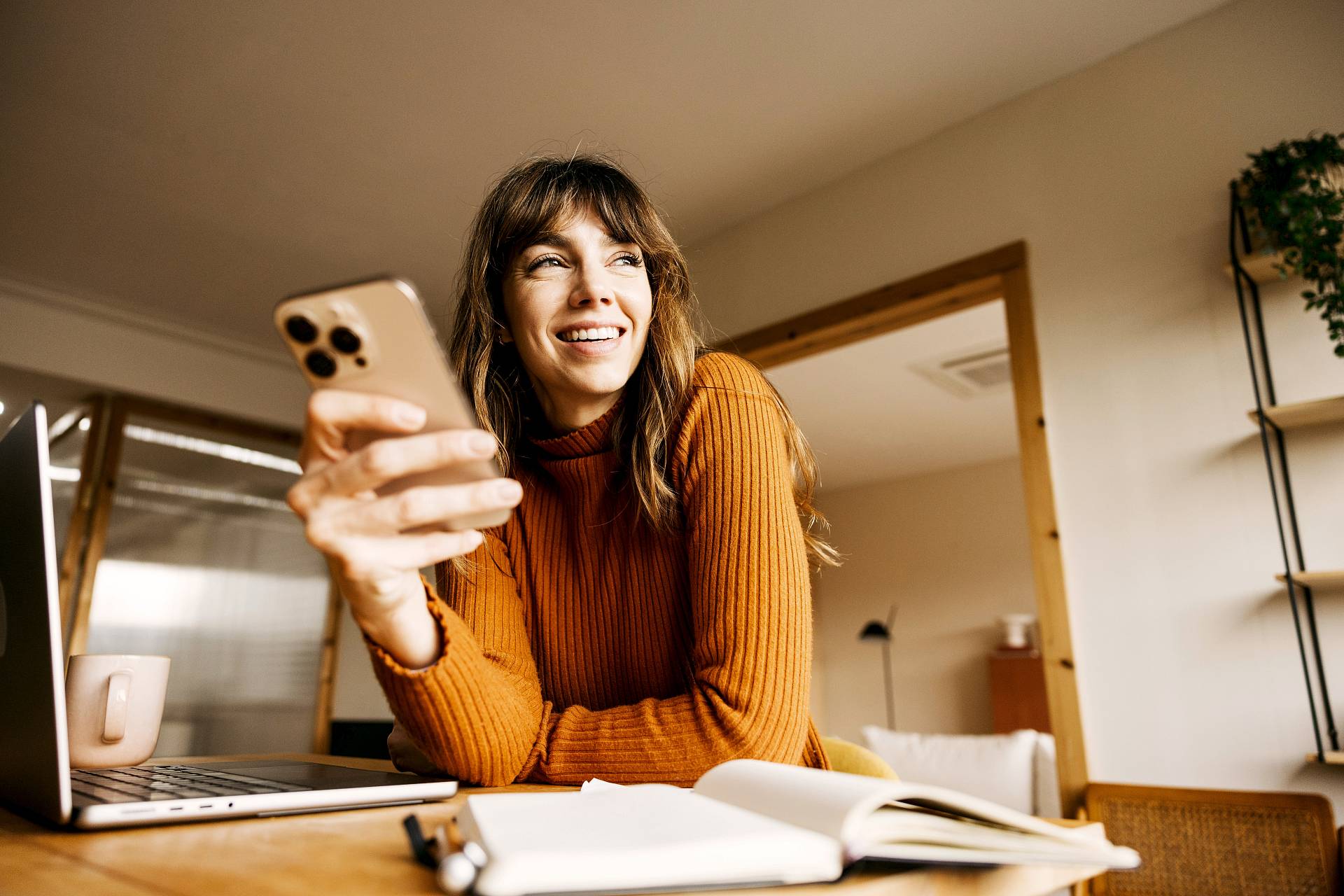 Lächelnde Frau sitzt mit Smartphone, Laptop und geöffnetem Notizbuch an einem Tisch.