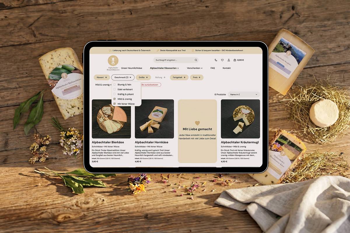 Tablet mit geöffnetem Käse-Onlineshop, umgeben von verschiedenen Käsesorten und Kräutern auf einem Holztisch.